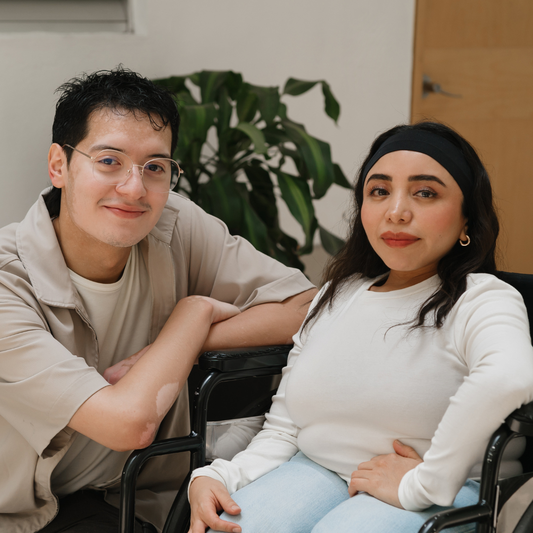 A young man kneels beside the wheelchair of a young woman. Both are smiling and look determined.