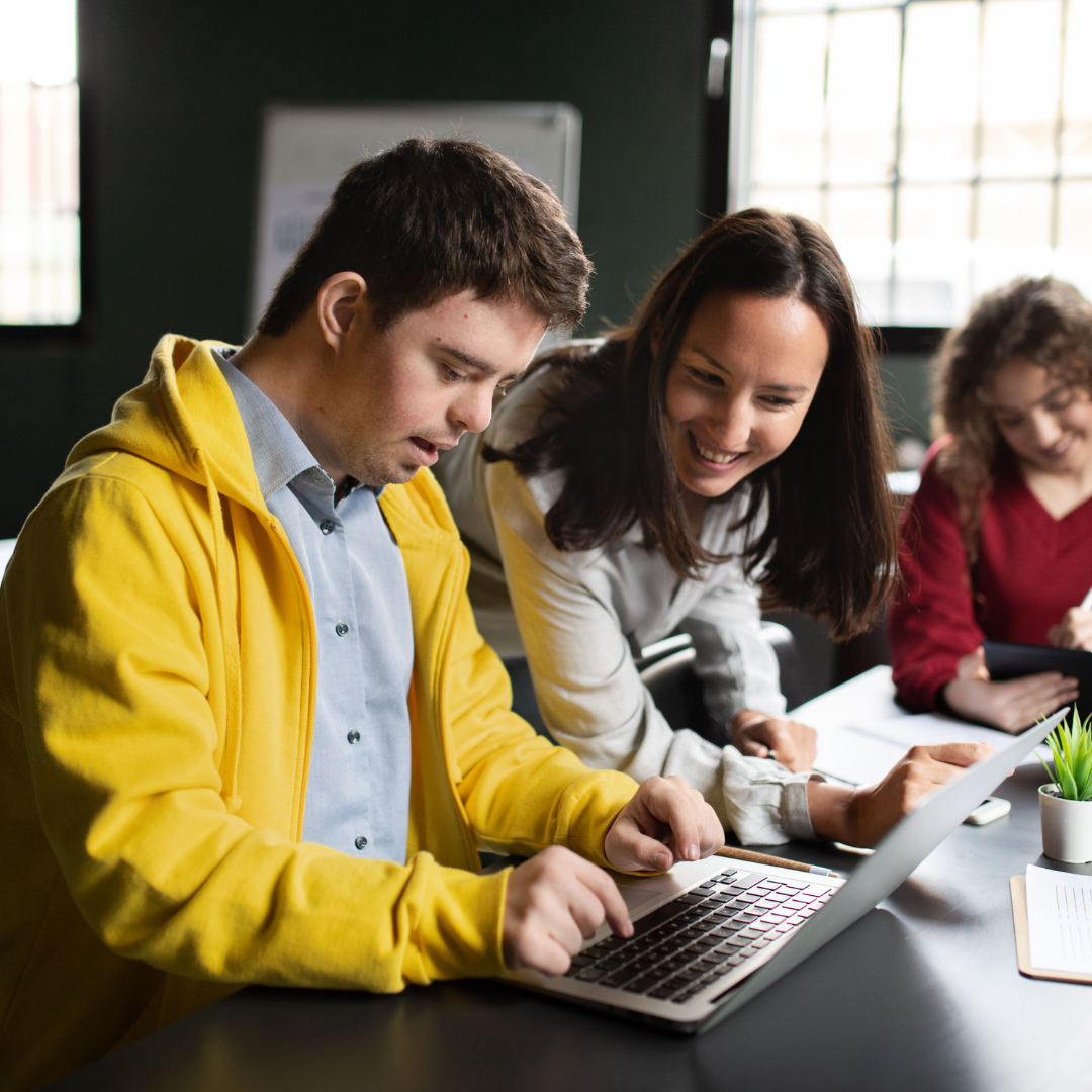 A young man with Down syndrome types on a computer. A woman is next to him looking at his screen and smiling.